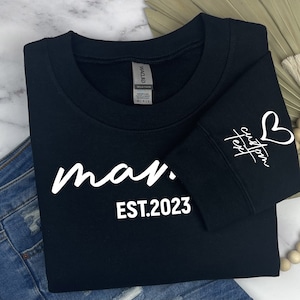 Op de afbeelding: Zwart sweatshirt met witte tekst die "man. EST.2023" luidt. De mouw heeft een wit hart en de tekst "custom text" in cursief.