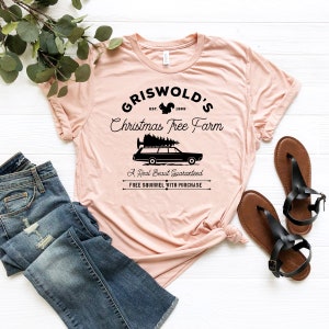 May include: A pink t-shirt with a black graphic design that says "Griswold's Christmas Tree Farm" with a squirrel, a car with a Christmas tree on top, and the text "A Real Beaut Guaranteed" and "Free Squirrel With Purchase".