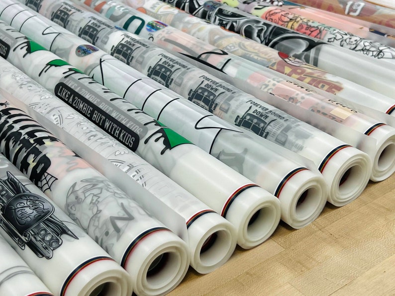 K&ouml;nnte beinhalten: Rollen aus klarem Vinyl mit verschiedenen bedruckten Designs, darunter ein LKW, ein Zombie und ein Sch&auml;del. Das Vinyl ist aufgerollt und hat einen roten, wei&szlig;en und schwarzen Streifen am Rand.