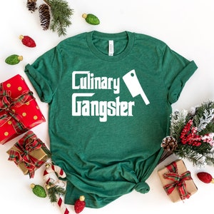 May include: Green t-shirt with white text that reads "Culinary Gangster" and a white silhouette of a butcher knife.