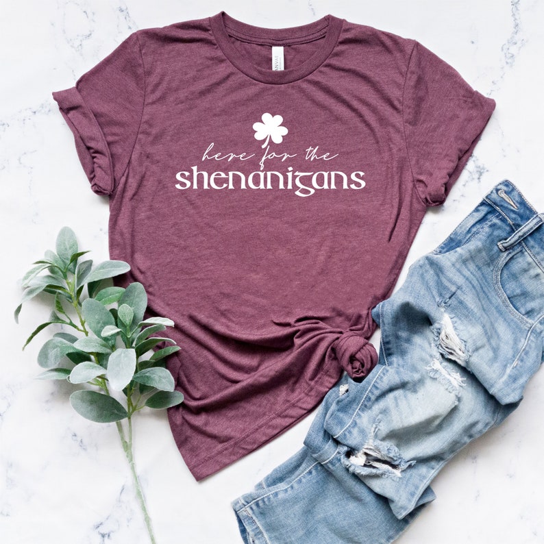 May include: A maroon t-shirt with a white shamrock and the text "here for the shenanigans".