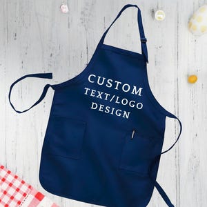 May include: A navy blue apron with a white pocket and adjustable straps. The apron has the text "CUSTOM TEXT/LOGO DESIGN" printed on the front.