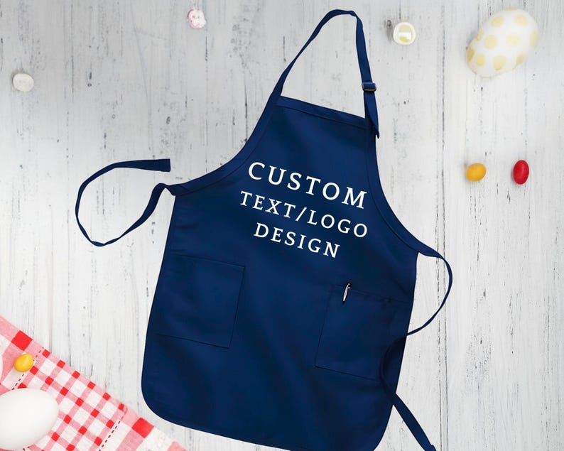 May include: A navy blue apron with a white pocket and adjustable straps. The apron has the text "CUSTOM TEXT/LOGO DESIGN" printed on the front.