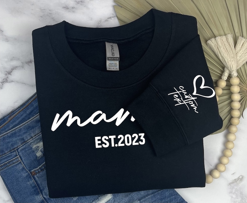 Op de afbeelding: Zwart sweatshirt met witte tekst die "man. EST.2023" luidt. De mouw heeft een wit hart en de tekst "custom text" in cursief.