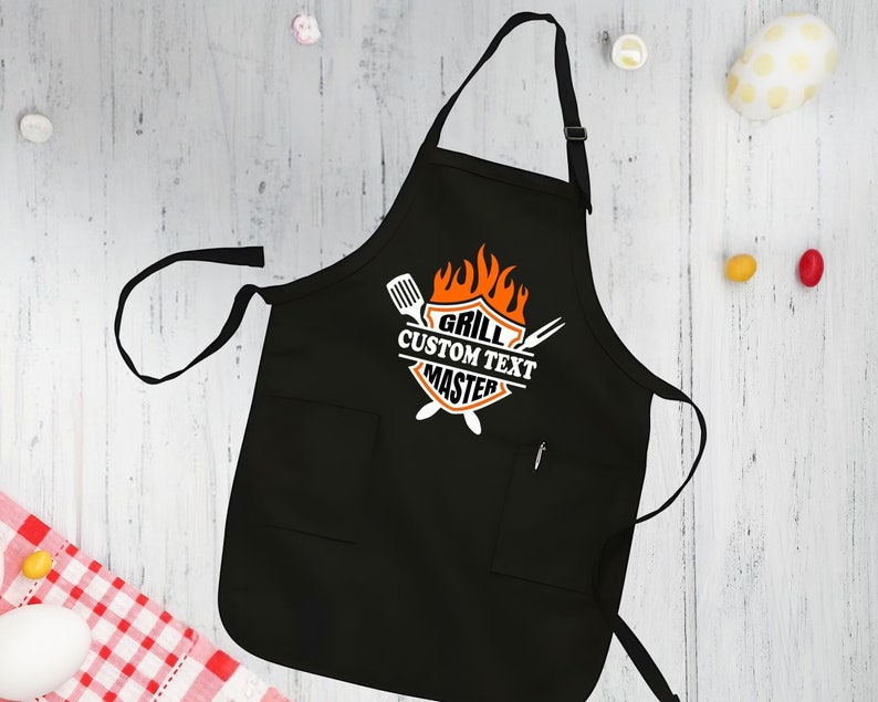 Puede incluir: Delantal de parrilla negro con un gr&aacute;fico blanco y naranja que dice "Grill Custom Text Master" con llamas y herramientas de parrilla.