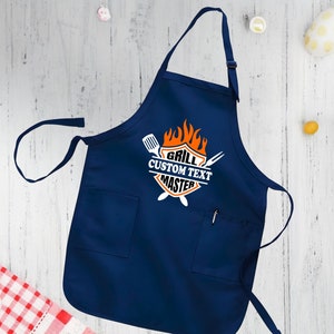 Puede incluir: Un delantal azul marino con un bolsillo blanco y un gr&aacute;fico blanco que dice "Grill Custom Text Master" con un dise&ntilde;o de llama y herramientas de parrilla cruzadas.