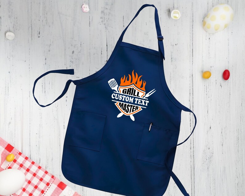 Puede incluir: Un delantal azul marino con un bolsillo blanco y un gr&aacute;fico blanco que dice "Grill Custom Text Master" con un dise&ntilde;o de llama y herramientas de parrilla cruzadas.