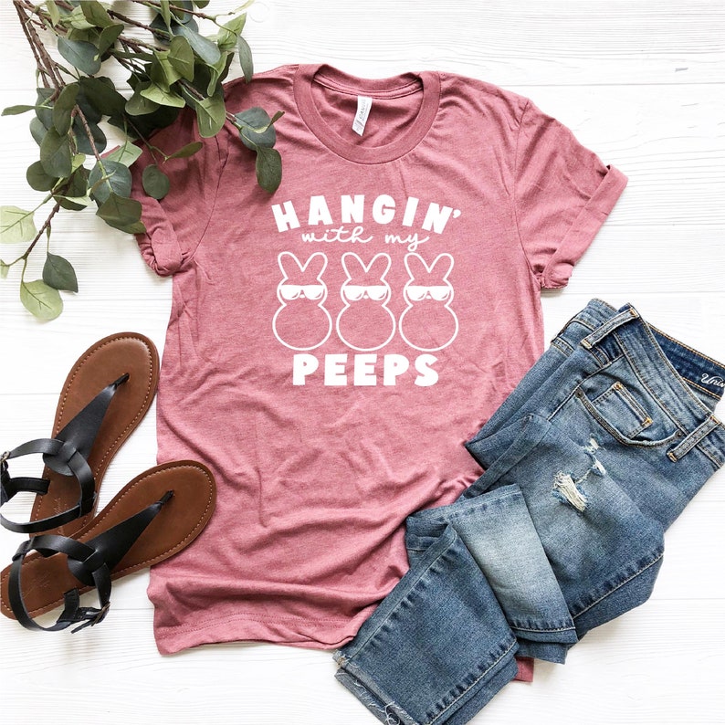 May include: A pink t-shirt with the text "Hangin' with my peeps" and three white cartoon peeps wearing sunglasses. The shirt is laid out on a white surface with a pair of blue jeans and sandals.