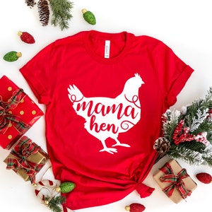 May include: Red t-shirt with a white silhouette of a chicken and the text "mama hen" on it. The shirt is laid out on a white surface with Christmas decorations around it.