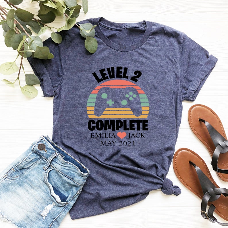 May include: A navy blue t-shirt with a retro sunset graphic and a video game controller. The text "Level 2 Complete" is printed on the shirt, along with the names "Emilia" and "Jack" and the date "May 2021".