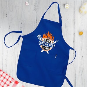 Puede incluir: Delantal de cocina azul con un gr&aacute;fico blanco y naranja que dice "Grill Custom Text Master" con un dise&ntilde;o de llama y herramientas de parrilla cruzadas.