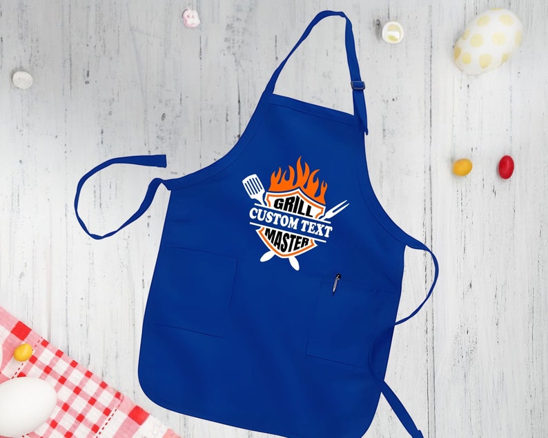 Puede incluir: Delantal de cocina azul con un gr&aacute;fico blanco y naranja que dice "Grill Custom Text Master" con un dise&ntilde;o de llama y herramientas de parrilla cruzadas.