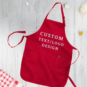 May include: Red apron with a large pocket and adjustable neck straps. The apron has white text that says "CUSTOM TEXT/LOGO DESIGN".