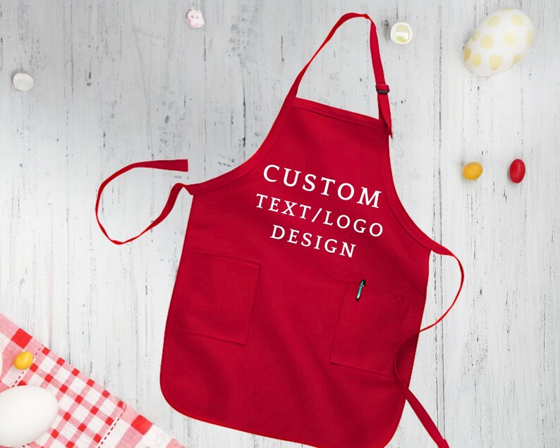May include: Red apron with a large pocket and adjustable neck straps. The apron has white text that says "CUSTOM TEXT/LOGO DESIGN".