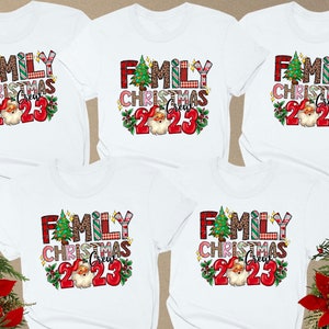 May include: White t-shirts with a colorful Christmas design. The design includes the text "FAMILY CHRISTMAS CREW 2023" with a Christmas tree, Santa Claus, and festive patterns. Red poinsettias and greenery frame the shirts.