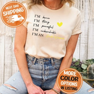 Pu&ograve; includere: Una maglietta beige con il testo "I'm brave, I'm strong, I'm powerful, I'm unbreakable, I'm a Cancer Warrior" in nero. Un cuore giallo &egrave; accanto al testo.