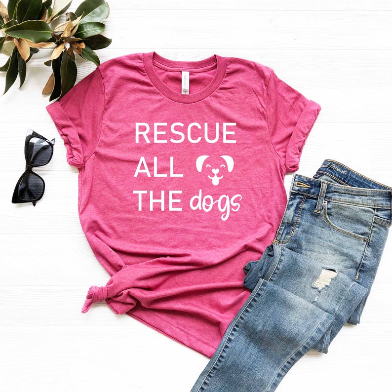 K&ouml;nnte beinhalten: Ein pinkfarbenes T-Shirt mit dem Text "RESCUE ALL THE dogs" in wei&szlig;en Buchstaben und einem wei&szlig;en Cartoon-Hund mit herausgestreckter Zunge.