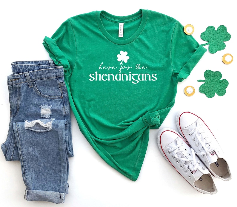 May include: Green t-shirt with a white shamrock and the text "Here for the shenanigans". The shirt is paired with blue jeans and white sneakers.