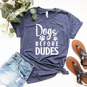 May include: A navy blue t-shirt with white text that reads "Dogs Before Dudes" with two paw prints above the word "Before".