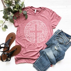 Puede incluir: Una camiseta rosa con un dise&ntilde;o gr&aacute;fico blanco de una cruz con las palabras "He is risen" y otras frases religiosas alrededor.