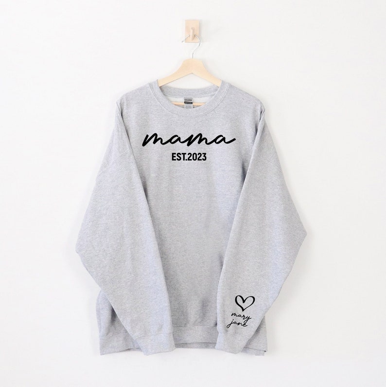 Op de afbeelding: Grijs sweatshirt met de tekst "mama est. 2023" op de voorkant en een hart met de tekst "mary jane" op de mouw.