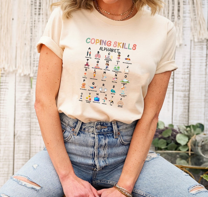 May include: A beige t-shirt with a graphic of a colorful alphabet chart that says "Coping Skills Alphabet". Each letter of the alphabet is paired with a coping skill, such as "Take a deep breath" for "B" and "Listen to music" for "M".