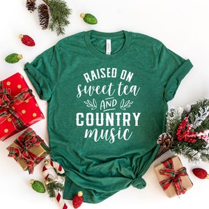 May include: Green t-shirt with white text that reads "Raised on sweet tea and country music". The shirt is photographed on a white background with Christmas decorations.