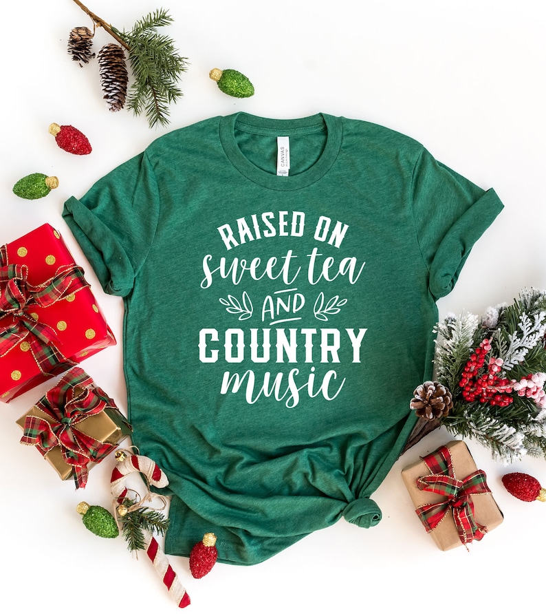 May include: Green t-shirt with white text that reads "Raised on sweet tea and country music". The shirt is photographed on a white background with Christmas decorations.