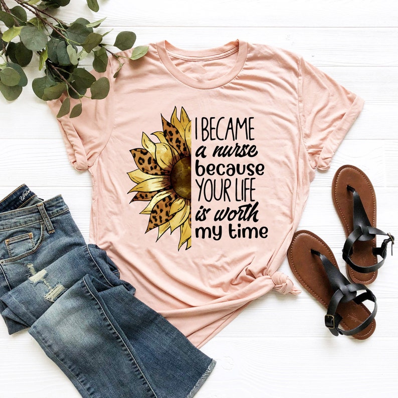 Op de afbeelding: Een roze T-shirt met een zonnebloemdessin en de tekst "Ik ben verpleegster geworden omdat je leven mijn tijd waard is".