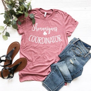 Puede incluir: Una camiseta rosa polvorienta con las palabras "Shenanigans Coordinator" en escritura blanca y un tr&eacute;bol. La camiseta se combina con vaqueros azules y sandalias negras, con una ramita de vegetaci&oacute;n.