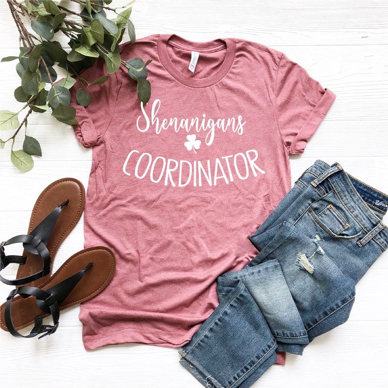 Puede incluir: Una camiseta rosa polvorienta con las palabras "Shenanigans Coordinator" en escritura blanca y un tr&eacute;bol. La camiseta se combina con vaqueros azules y sandalias negras, con una ramita de vegetaci&oacute;n.