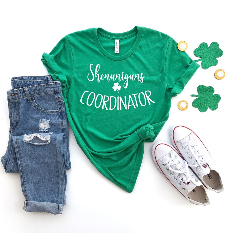 Puede incluir: Una camiseta verde con la frase "Shenanigans Coordinator" en blanco, combinada con vaqueros azules y zapatillas blancas. La imagen incluye decoraciones de tr&eacute;boles y monedas, ideal para el D&iacute;a de San Patricio.