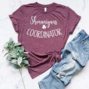 Puede incluir: Una camiseta granate con las palabras "Shenanigans COORDINATOR" en escritura blanca y un tr&eacute;bol. Se combina con vaqueros de mezclilla azul claro y desgastados. Una ramita de hojas verdes a la izquierda.