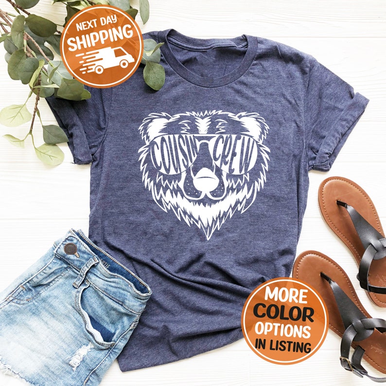 May include: A navy blue t-shirt featuring a white bear graphic wearing sunglasses. The sunglasses have the words "Cousin Crew" written on them.