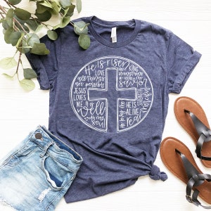 Puede incluir: Una camiseta azul marino con un gr&aacute;fico blanco de una cruz rodeada de palabras, incluyendo "He is risen", "Love", "Jesus loves me", "It is well", "He strengthens my friend me", "Savior", "Strength", "Merciful", "He is alive", y "Real".