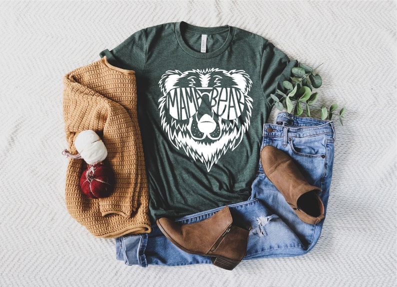 May include: A green t-shirt with a white bear graphic wearing sunglasses. The text "Mama Bear" is inside the sunglasses. The shirt is laid out on a white surface with a brown sweater, blue jeans, and brown boots.