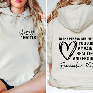 May include: A cream-colored hoodie with a black heart and the text "You Matter" on the front. The back of the hoodie has a black heart and the text "To the person behind me: You are amazing, beautiful and enough. Remember That."