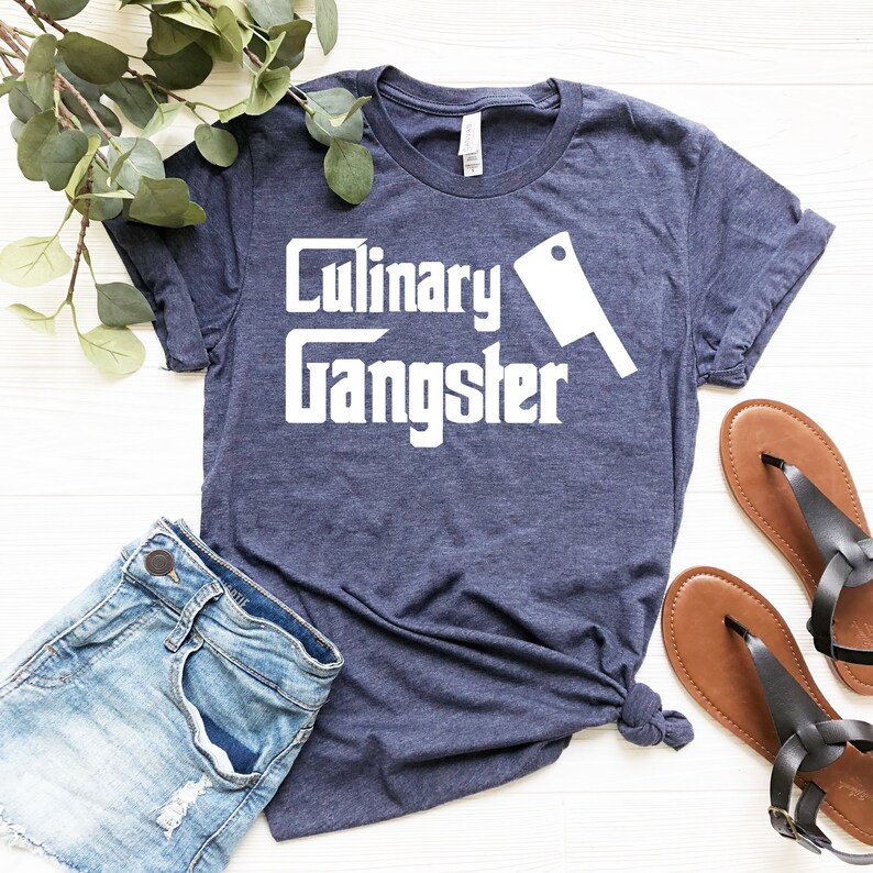 May include: A navy blue t-shirt with the text "Culinary Gangster" in white lettering and a white silhouette of a butcher knife.