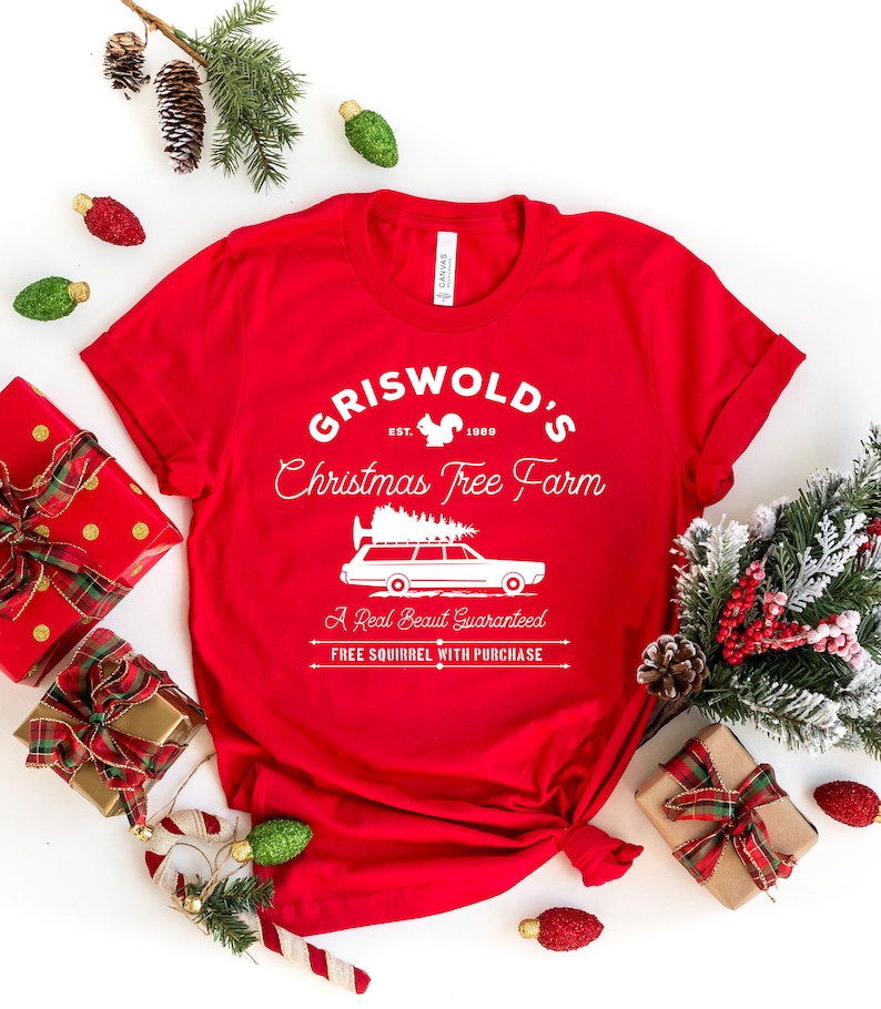 May include: Red t-shirt with a white graphic design that says "Griswold's Christmas Tree Farm" with a squirrel, the year 1989, and a cartoon car with a Christmas tree on top. The text below the graphic says "A Real Beaut Guaranteed" and "FREE SQUIRREL WITH PURCHASE."