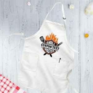 Puede incluir: Delantal blanco con un logotipo de maestro de la parrilla naranja y negro. El logotipo presenta una parrilla, llamas y el texto "Grill Custom Text Master".