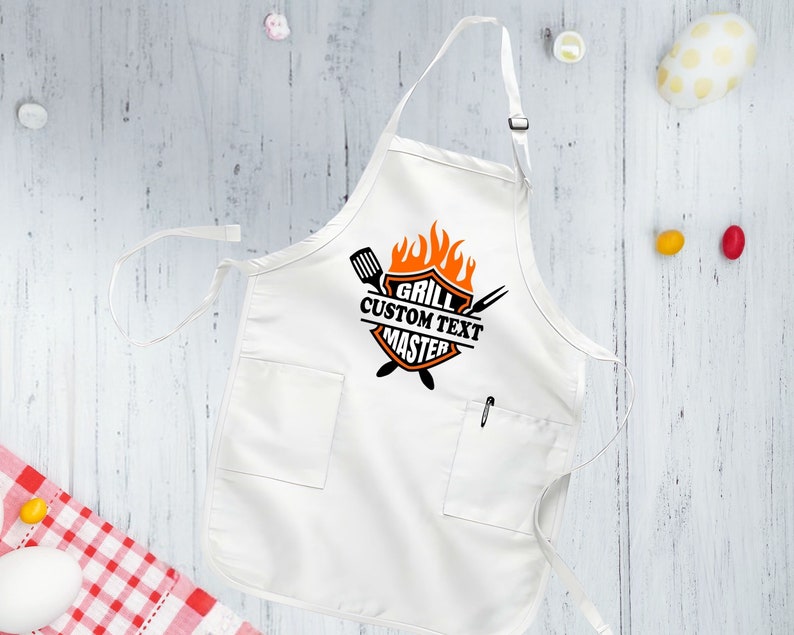 Puede incluir: Delantal blanco con un logotipo de maestro de la parrilla naranja y negro. El logotipo presenta una parrilla, llamas y el texto "Grill Custom Text Master".