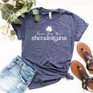 May include: A navy blue t-shirt with a white shamrock and the text "here for the shenanigans".