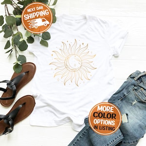 May include: White t-shirt with a gold sun and moon design. The sun has rays and the moon is a crescent shape. There are small stars around the sun and moon.