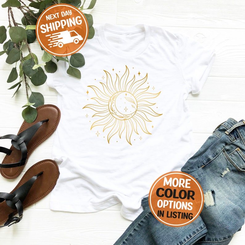 May include: White t-shirt with a gold sun and moon design. The sun has rays and the moon is a crescent shape. There are small stars around the sun and moon.