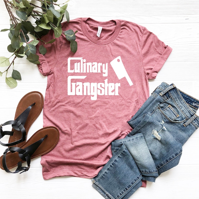 May include: A heather pink t-shirt with the text "Culinary Gangster" in white, featuring a white chef's knife graphic. The shirt is laid flat on a white surface with a pair of black sandals and blue jeans.