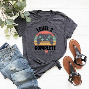 May include: A gray t-shirt with a retro-style graphic of a video game controller and the text "Level 2 Complete" with the names "Emilia" and "Jack" and the date "May 2021".