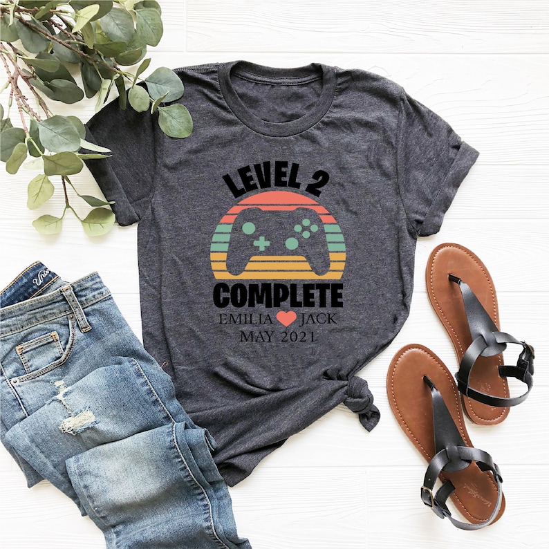 May include: A gray t-shirt with a retro-style graphic of a video game controller and the text "Level 2 Complete" with the names "Emilia" and "Jack" and the date "May 2021".