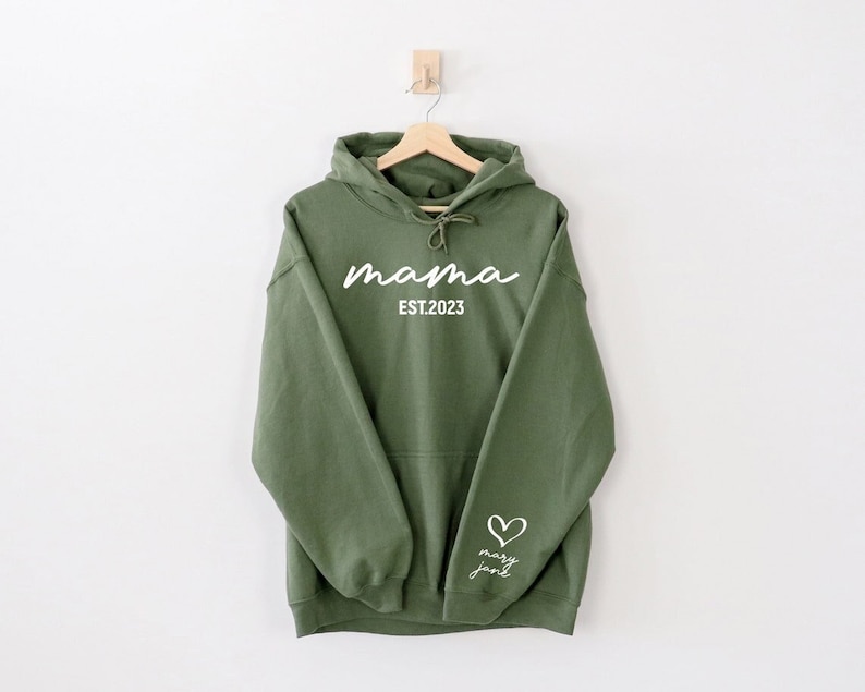 Op de afbeelding: Een groene hoodie met de tekst "mama est. 2023" op de voorkant en een hart met de tekst "mary jane" op de mouw.