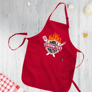 Puede incluir: Delantal de cocina rojo con un bolsillo blanco y un gr&aacute;fico que dice "Grill Custom Text Master" con un dise&ntilde;o de llama y utensilios de parrilla cruzados.