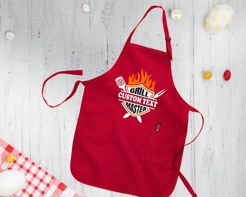 Puede incluir: Delantal de cocina rojo con un bolsillo blanco y un gr&aacute;fico que dice "Grill Custom Text Master" con un dise&ntilde;o de llama y utensilios de parrilla cruzados.